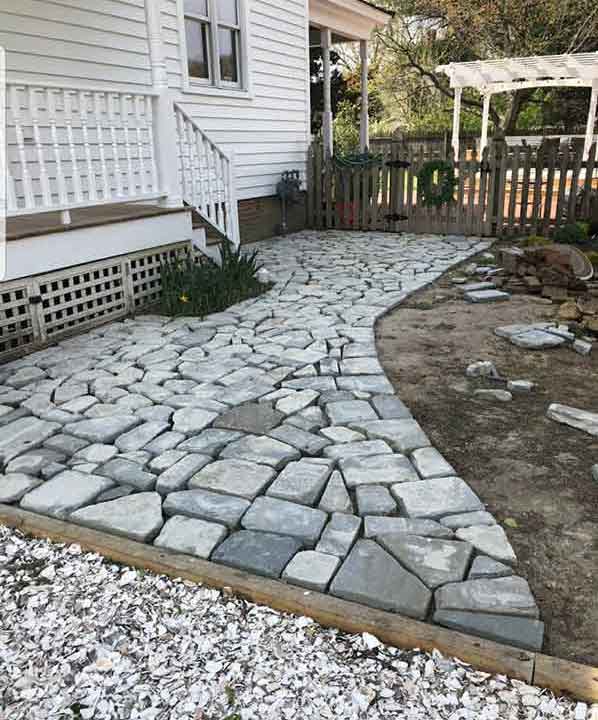 A nice stone walk coming from a gravel driveway makes a nice transition and is flanked by a wood stairwell with small porch and a wood barrier.
